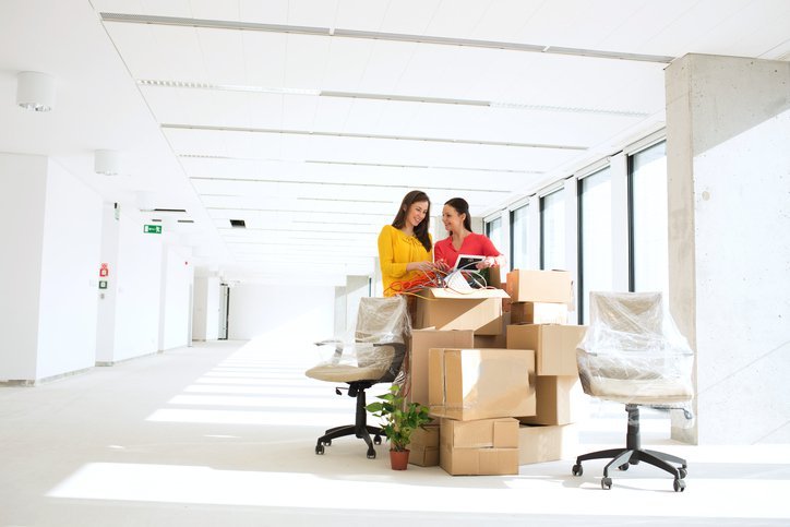 Two women discussing office move preparations surrounded by stacked moving boxes in a bright, empty office space, emphasizing the theme of office move-out cleaning services in Melbourne.
